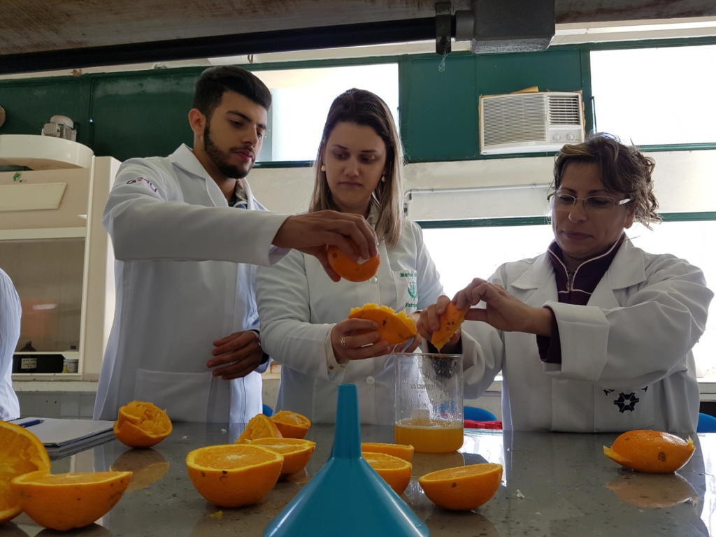 Três pessoas vestindo jaleco branco trabalhando com laranjas em uma bancada