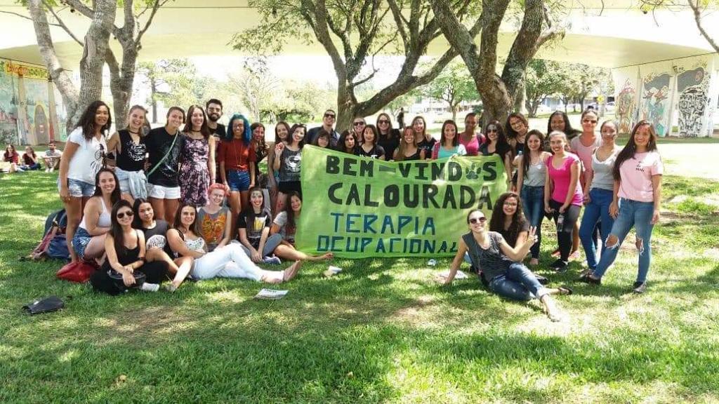 Turma de estudantes em um gramado segurando faixa verde escrito Recepção calouros Terapia Ocupacional