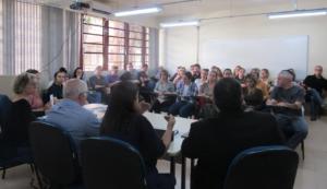 Fotografia horizontal tirada do fundo para a frente da sala. Dois homens e duas mulheres compõe a mesa de palestrantes e estão voltados para um grupo de pessoas sentadas a frente.