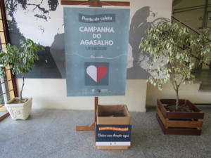 fotografia horizontal e colorida de um banner em um suporte de madeira, a frente uma caixa de papelão e nas laterais dois vasos de plantas, um em cada lado da imagem.